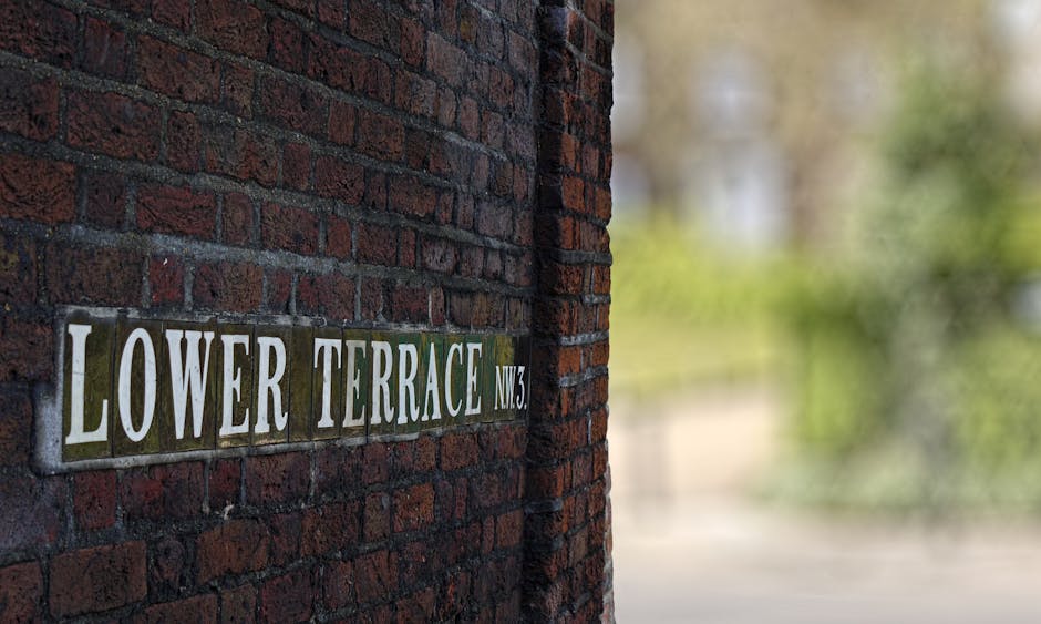 Close-up view of a brick wall displaying a rectangular metal street sign with white lettering that reads 'Lower Terrace NW3', mounted on a dark red brick exterior wall. The background is blurred and shows a soft focus of greenery and possibly a sidewalk or street, indicating an outdoor residential or urban environment. The sign is positioned to the left of the image, with the wall extending vertically. The scene suggests a location within a neighbourhood, relevant to house removals and home relocation services provided by Man with Van Belsize Park, focusing on outside property surroundings and street identification for moving logistics.